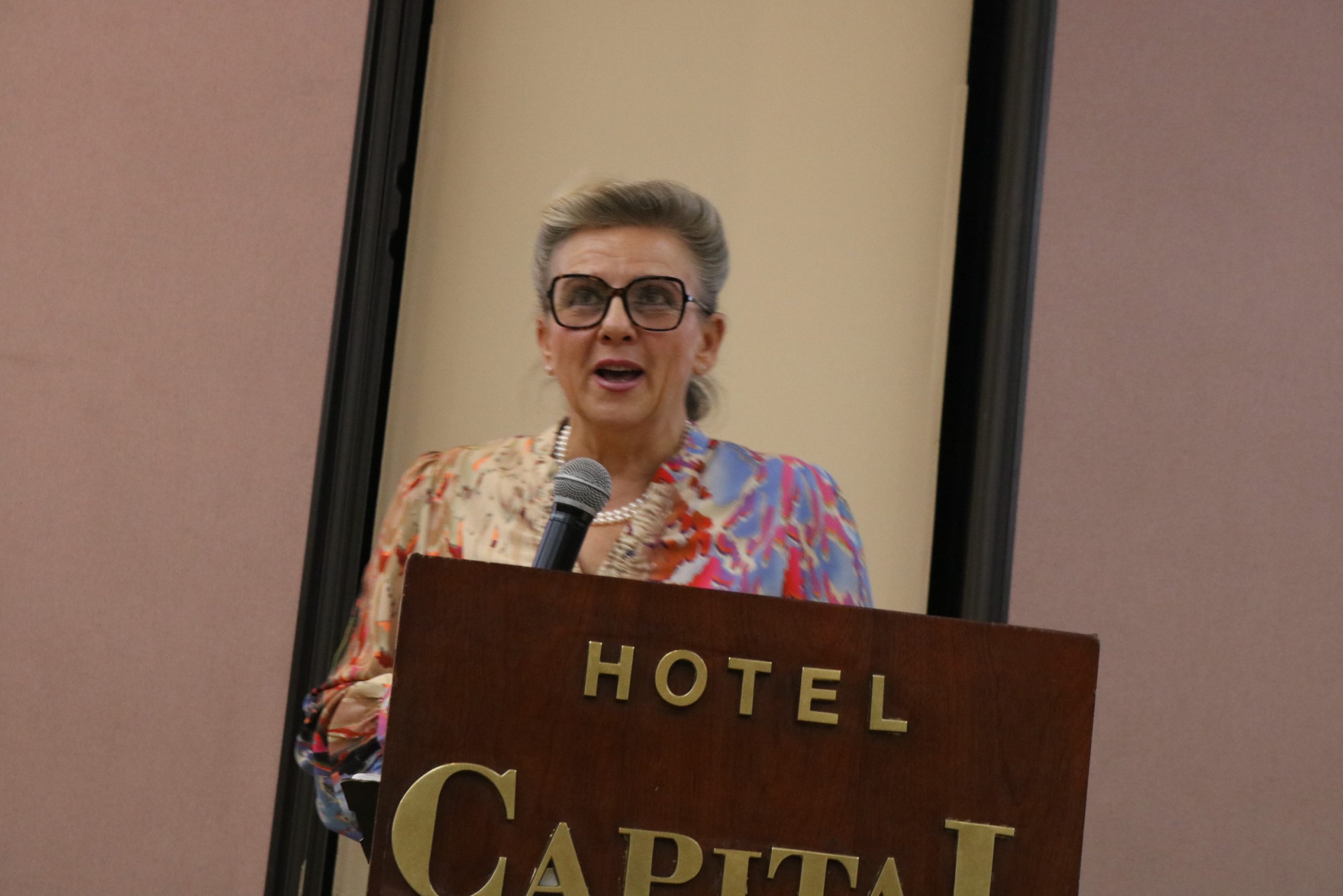 Mujer adulta con gafas y blusa colorida habla en un podio del Hotel Capital sosteniendo un micrófono.