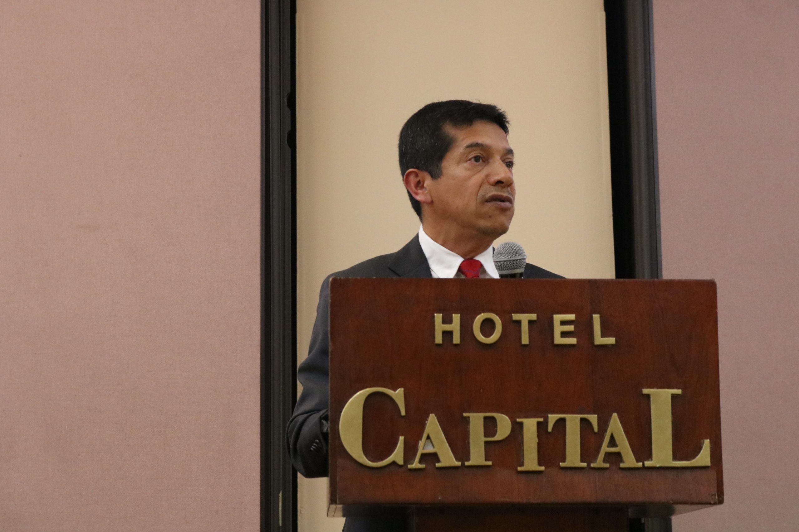 Hombre con traje oscuro y corbata roja habla en un podio de madera del Hotel Capital sosteniendo un micrófono.